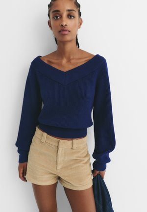 Navyblauwe gebreide trui met ruime V-hals en lange mouwen, gecombineerd met beige corduroy korte broek met voorzakken en een soepele textuur.