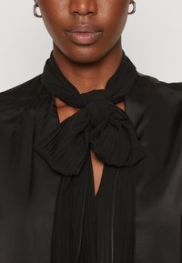 Blusa nera con un nodo al collo, plissettata, e una texture satinata. Il tessuto ha una finitura liscia con ritagli vicino al colletto.
