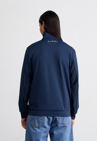Marinska zip-up jakna z visokim ovratnikom, prilegajočimi manšetami in teksturirano blago. Ima subtilen logotip blizu ovratnika. Obrabljena z svetlo modrimi kavbojkami.