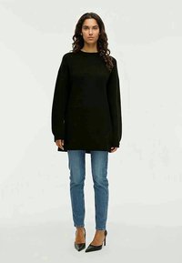 Nainen, jolla on kihara hiukset, pukeutuneena mustaan ylipitkään neuleeseen, sinisiin kapeisiin farkkuihin ja mustiin korkokenkiin, seisoo yksinkertaista taustaa vasten.