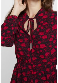 Robe noire avec un motif floral rouge, dotée d'un col en trou de clé, d'un détail noué et d'une taille élastique. Manches longues avec des poignets froncés.