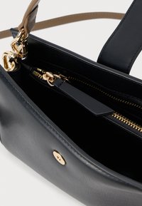 Melnas ādas rokassomas iekšpuse ar zelta toņu detaļām, tajā ir rāvējslēdzēja kabata un atsevišķa plakana kabata, gluda tekstūra un minimālistisks dizains.