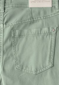 Tela de mezclilla verde menta con un bolsillo trasero que presenta un contorno cosido y un detalle de botón de metal. La etiqueta dice "STREET ONE DENIM ATELIER."