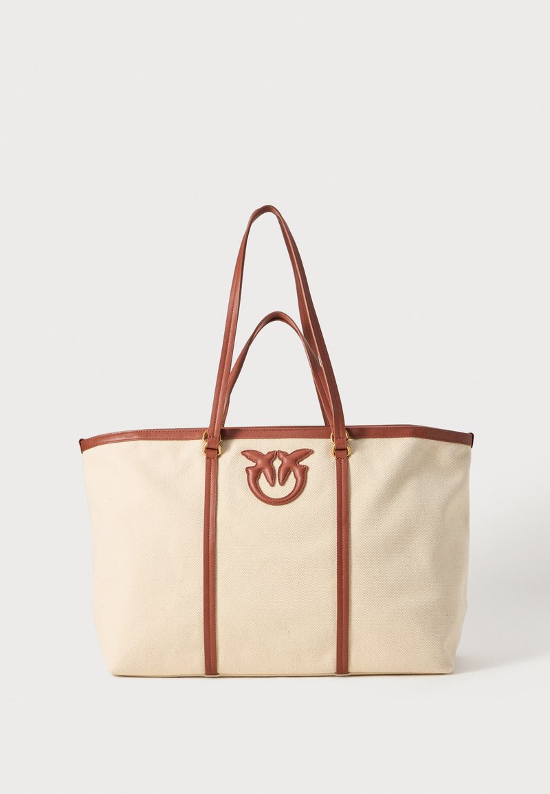 Beige bomullsveske med brune lærhåndtak og detaljer, med en fremtredende preget logo på forsiden. To vertikale lærakenter.