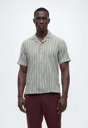 Mann steht vor neutralem Hintergrund und trägt ein kurzärmeliges, grau gestreiftes Hemd mit Knöpfen sowie dunkelrote Hosen.