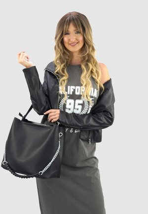 Jonge vrouw met golvend blond haar draagt een zwarte leren jas over een grijze California 95 jurk en houdt een grote zwarte handtas met zilveren ketting vast.