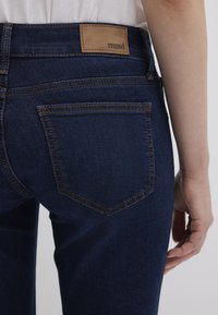 Dark blue denim jeans with a smooth texture, featuring a leather tag with "mavi" on the waistband and contrasting stitching on the pockets.