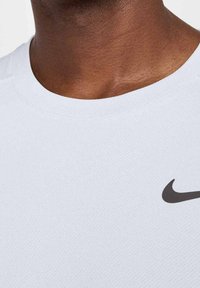 Camiseta deportiva blanca con una tela texturada, que presenta un logo de Nike Swoosh negro en la parte frontal y un cuello redondo.