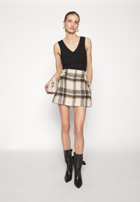 Crna majica bez rukava s V izrezom, u kombinaciji s tartan mini suknjom u krem, sivoj i smeđoj boji. Manekenka drži krem torbu i nosi crne gležnjače.