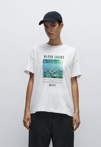 Hvid bomulds t-shirt med et trykt Monet kunstværk tituleret "Vandliljer", som viser livlige grønne og blå nuancer med sort tekst nedenfor.