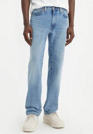 Personne portant un jean droit bleu clair et des chaussures décontractées ivoire, debout devant un fond uni.