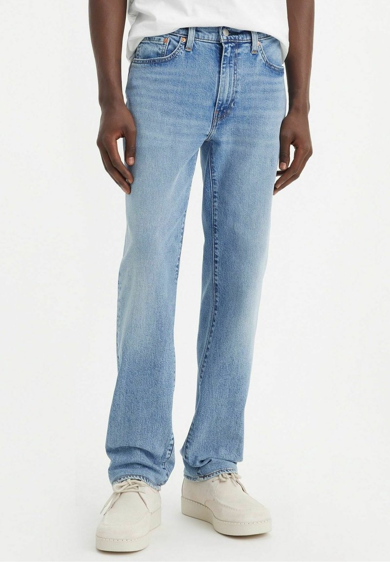Personne portant un jean droit bleu clair et des chaussures décontractées ivoire, debout devant un fond uni.