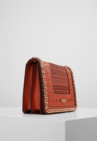 Czerwona torebka z eko skóry z ozdobnym wzorem wycięć, złotym łańcuszkiem przy krawędziach i prostokątnym, usztywnionym kształtem.