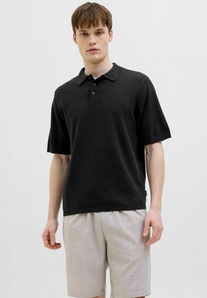 Jeune homme portant un polo noir à manches courtes et un short beige clair, debout devant un fond uni gris clair.
