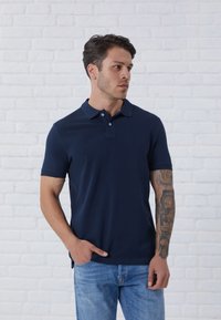 Navy blue polo shirt with short sleeves, a three-button placket, and side slits, worn with light blue jeans against a white brick wall.