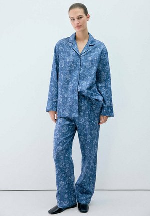 Jeune femme portant un haut de pyjama bleu à fleurs à manches longues boutonné et un pantalon ample assorti avec des chaussures noires, debout devant un fond blanc uni.