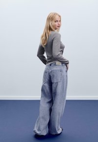 Grauer Langarmoberteil kombiniert mit hellblauen weiten Jeans. Die Jeans verfügen über zwei Gesäßtaschen und ein helles Taillenetikett.