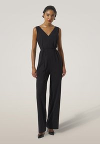 Kvinna som står klädd i en ärmlös svart jumpsuit med V-ringning och knytbälte, i kombination med svarta högklackade skor mot en ljus bakgrund.