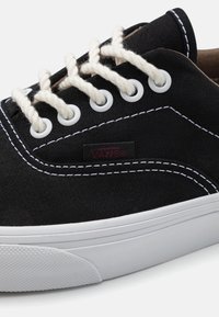 Czarny płócienny trampek z białym przeszyciem, okrągłymi białymi oczkami, jasnymi sznurówkami i teksturowaną białą gumową podeszwą. Na bucie znajduje się metka z logo Vans.