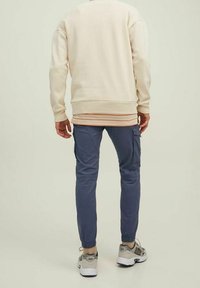 Osoba stojící, oblečená v krémovém svetru, pod kterým má pruhovanou košili, modrých cargo teplákových kalhotách a béžových teniskách na jednolitém pozadí.