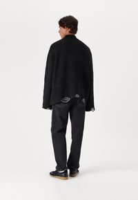 Μαύρος υπερμεγέθης πλεκτός σκούφος με ακατέργαστες άκρες, φορεμένος πάνω από σκούρα τζιν και μαύρα αθλητικά παπούτσια με καφέ σόλες, απεικονισμένος από πίσω.