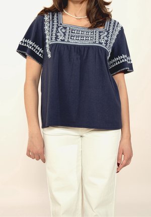 Top de algodón azul marino de manga corta con escote cuadrado y detalles bordados intrincados en blanco en las mangas y el cuerpo. Combinado con pantalones blancos.