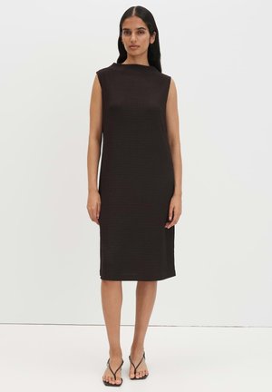 Frau steht, trägt ein ärmelloses schwarzes knielanges Kleid und schwarze Riemchensandalen vor einem schlichten hellen Hintergrund.