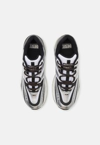 Sneakers med hvit mesh-overdel, svarte detaljer og brune snørebånd; har en rund gummisåle og teksturerte designelementer på sidene.