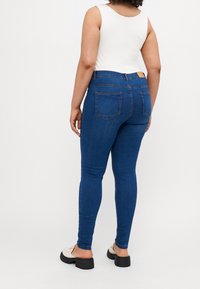 Jeans skinny de denim azul oscuro con cinco bolsillos, cintura alta y un ligero estiramiento. Textura suave, sin patrones visibles y con un hardware mínimo.