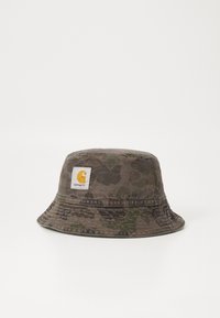 Bucket hat i camouflage-mønster; stoffet er blødt bomuldsstof. Har en syet skygge og et firkantet logo-patch i gul og hvid på forsiden.