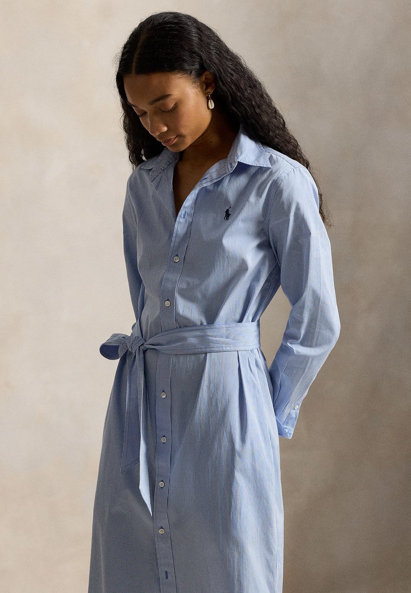 Φόρεμα πουκάμισο με κουμπιά, μπλε και λευκές ρίγες, γιακά, ζώνη στη μέση, μακριά μανίκια και μικρό λογότυπο στο στήθος. Λείο υλικό υφής.