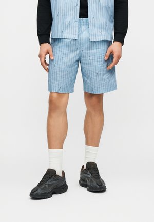 Mężczyzna w jasnoniebieskich szortach w prążki i dopasowanej otwartej koszuli na czarną bluzę z długim rękawem, białe skarpetki i czarne trampki.