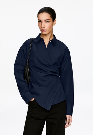 Femme aux cheveux tirés en arrière portant une chemise portefeuille bleu marine et un pantalon noir, portant un sac à bandoulière noir, debout devant un fond blanc uni.
