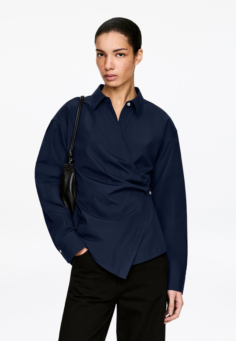 Femme aux cheveux tirés en arrière portant une chemise portefeuille bleu marine et un pantalon noir, portant un sac à bandoulière noir, debout devant un fond blanc uni.