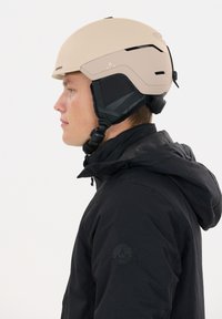 Beiger Skihelm mit glatter, matter Oberfläche, integriertem Ohrenschutz, verstellbaren Riemen und einer schwarzen Jacke mit einem runden Logo.