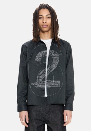 Person mit langen Dreadlocks, der ein schwarzes Hemd mit dünnen Streifen und einer großen weißen Zahl 2 über einem weißen T-Shirt trägt.