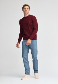 Burgundy stickad tröja med kabelmönster, i kombination med ljusblå jeans och beige sneakers. Modellen står framför en neutral bakgrund.