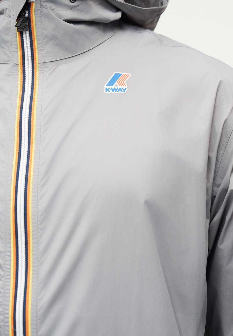 Veste gris clair avec une fermeture éclair rayée jaune, blanche et noire, arborant un logo K-Way bleu et orange sur la poitrine.