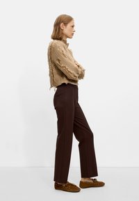 Beżowy sweter dziany z postrzępionymi krawędziami, brązowe spodnie o dopasowanym kroju oraz brązowe buty typu mokasyny z ozdobnymi sznurówkami. Widok z profilu bocznego.