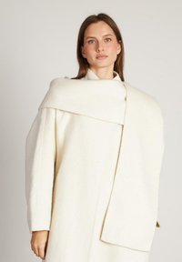 Cappotto bianco in lana con un design strutturato e ampio colletto. Le caratteristiche principali includono maniche lunghe e un dettaglio drappeggiato. Texture liscia.