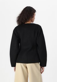 Svart ullblazer med strukturerte skuldre, sømdetaljer og en tettsittende silhuett, sammen med beige vide bukser.