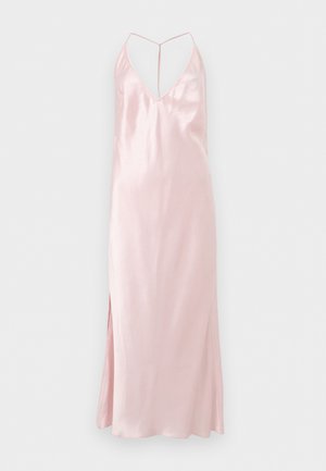 Frau trägt ein hellrosa Satin-Nachthemd mit Spaghettiträgern und schwarze Riemchen-High-Heel-Sandalen, steht mit den Händen hinter dem Rücken.