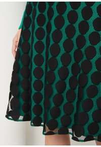 Falda verde con grandes lunares negros y sobrecapa de malla, mostrada desde medio muslo hasta mano descansando a un lado.