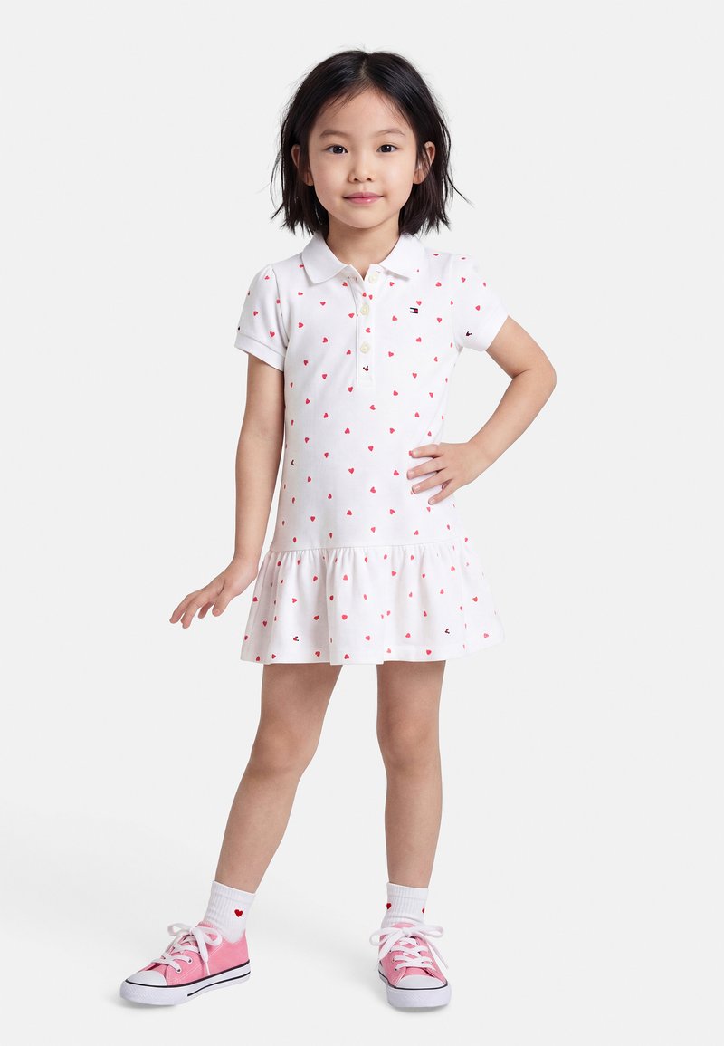 Νεαρή κοπέλα står με το χέρι στη μέση, φορώντας λευκό φόρεμα με κόκκινα καρδιές, λευκές κάλτσες και ροζ αθλητικά παπούτσια σε λευκό φόντο.