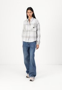 Camisa de botão xadrez em cinza e branco, com dois bolsos no peito e um logótipo em patch. Combinada com jeans de perna larga azuis e ténis.