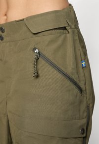 Olijfgroene cargobroek met schuine ritszak, klepzak met drukknoop, verstelbare tailleband en een klein Zweeds vlaggetje aan de zijkant.