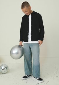 Schwarzes Button-Up-Hemd, darunter ein weißes T-Shirt, hellblaue weit geschnittene Jeans und schwarze Sneakers. Hält einen silbernen Ballon.