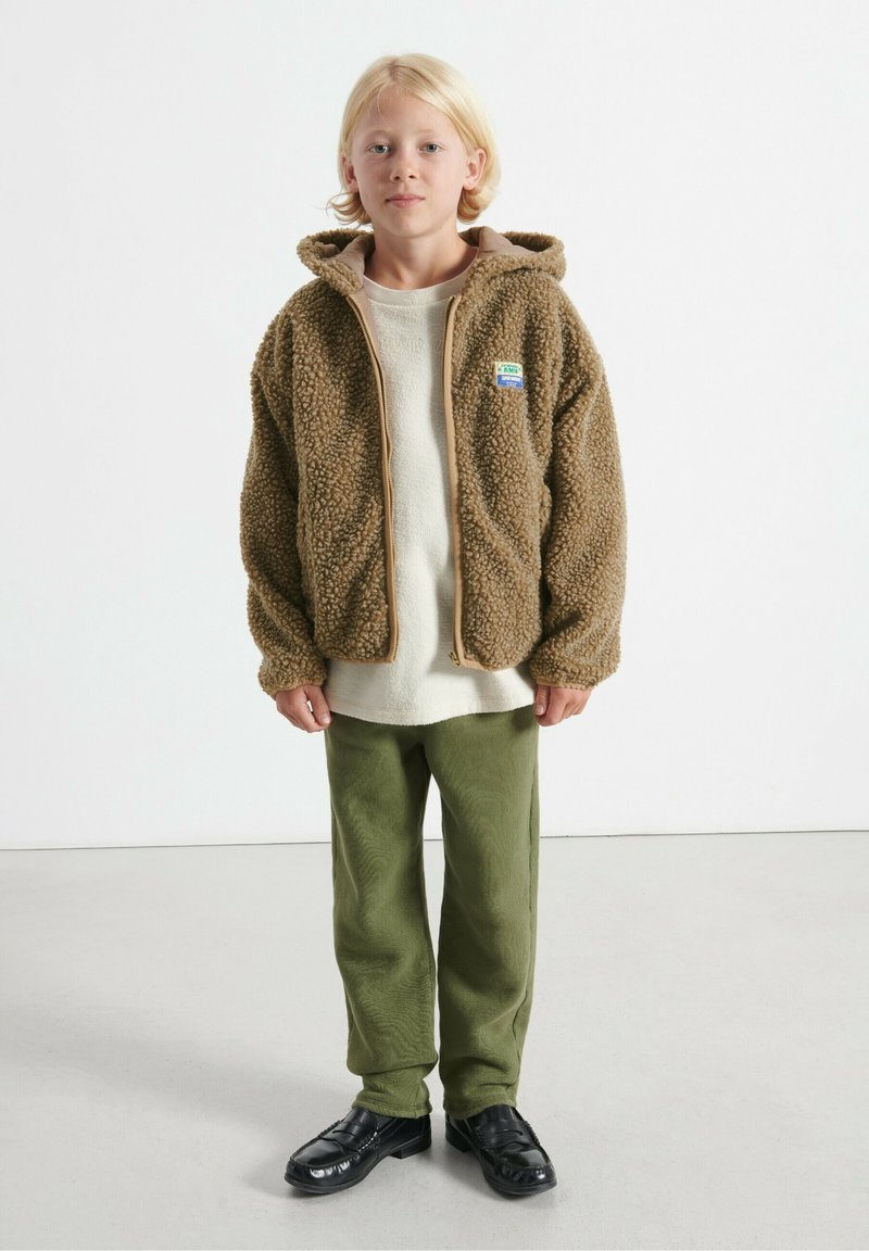 Bambino biondo in piedi, con felpa a zip in pile marrone, maglietta crema, pantaloni verdi e scarpe nere senza lacci, su uno sfondo bianco semplice.