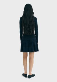 Neulemekko tummansinisena, pitkät hihat, istuva vartalo ja laskostettu hame. Yhdistetty mustiin tasapohjaisiin kenkiin; hiukset ulottuvat olkapäiden yli.