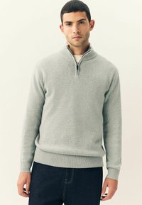 Hombre con un suéter de media cremallera gris claro y jeans oscuros, frente a un fondo liso claro, mirando hacia adelante con una expresión neutral.